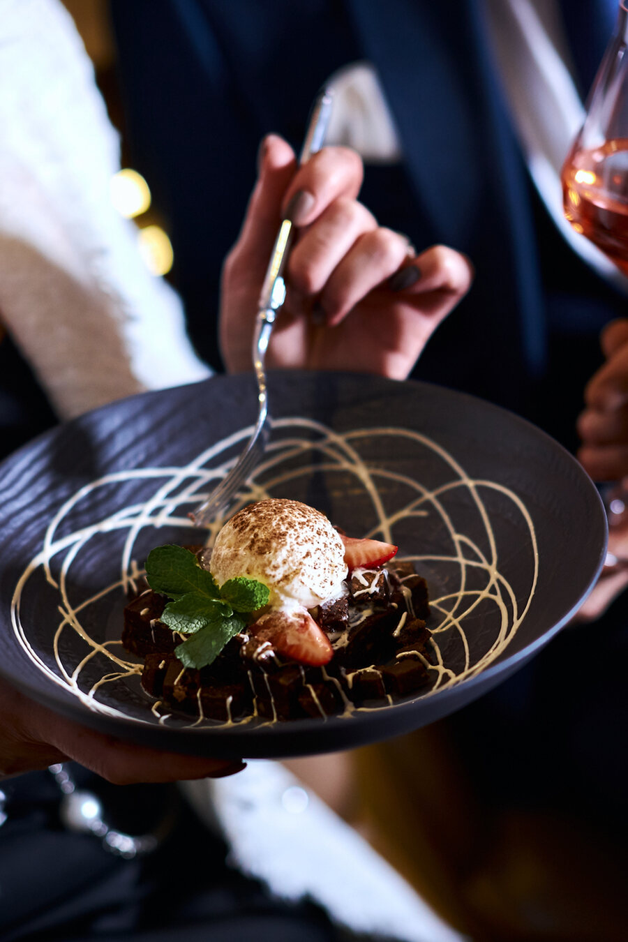 Hände servieren am Gasttisch einen Dessertteller mit Minze und Erdbeere, daneben ein Glas Roséwein