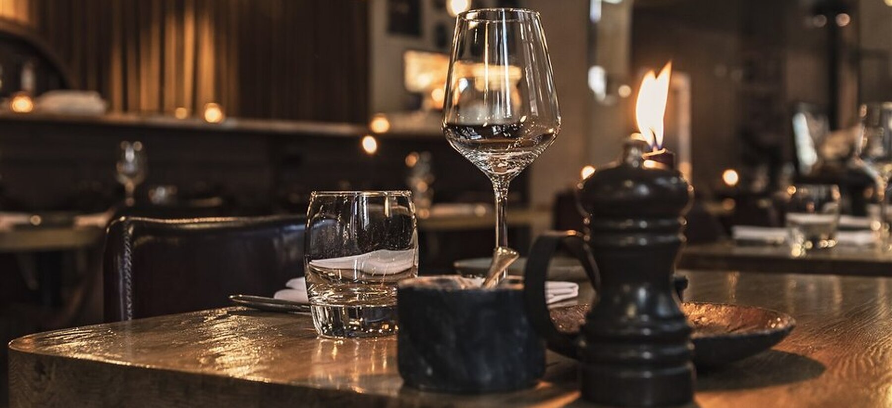 Weinglas, Wasserglas und brennende Tischleuchte auf einem Holztisch im Gastraum am Abend