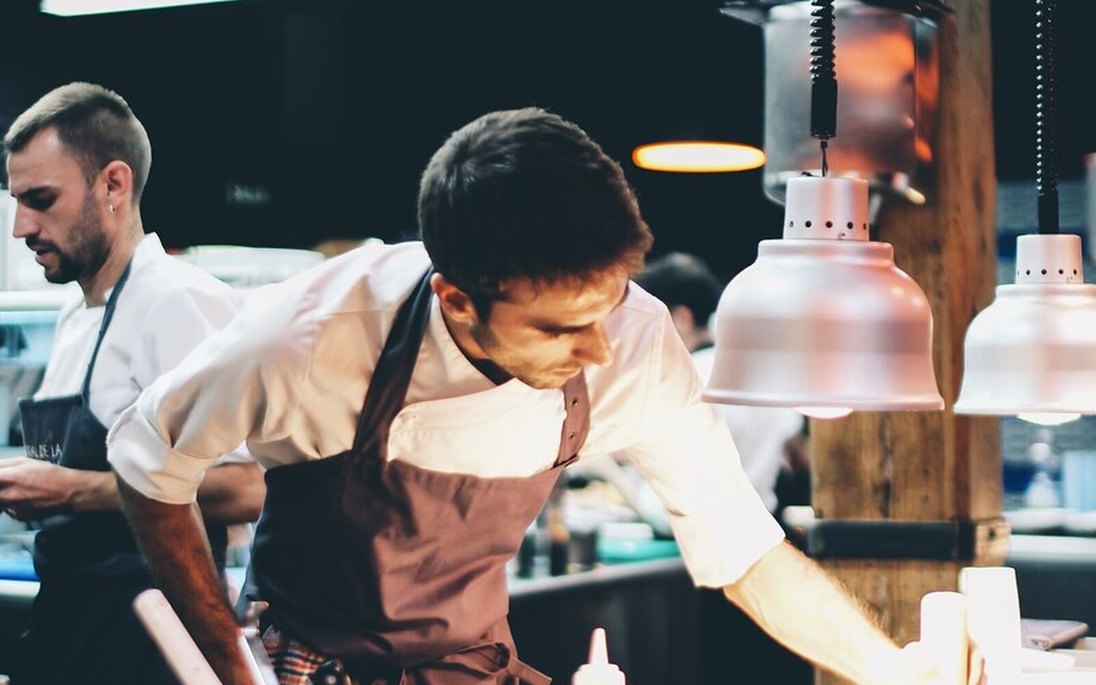 Zwei Köche richten am Pass einer Restaurantküche unter Wärmelampen an — Arbeitsalltag in einem Haus, das die Werbeagentur für Gastronomie betreut